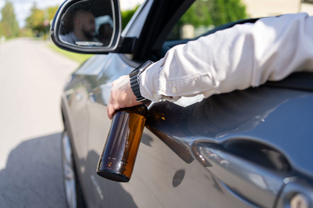 A drunken man driving a car with a bottle of alcohol in his hand. Close-up of a bottle of Alcohol in the driver hand at the wheel of car. Alcohol driving concept.